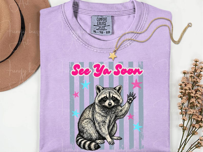 Peut inclure: T-shirt lilas avec un dessin de raton laveur qui salue et le texte "See Ya Soon". Le motif comprend des &eacute;toiles roses et bleues et des rayures verticales grises. Un collier dor&eacute; avec un pendentif en forme d'&eacute;toile est pos&eacute; sur le t-shirt.