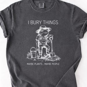 May include: Dark gray t-shirt with a white graphic of a frog in overalls and a hat, holding a shovel. Text reads "I BURY THINGS... MAYBE PLANTS... MAYBE PEOPLE."