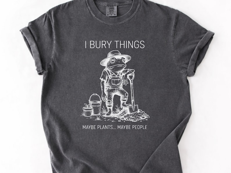 May include: Dark gray t-shirt with a white graphic of a frog in overalls and a hat, holding a shovel. Text reads "I BURY THINGS... MAYBE PLANTS... MAYBE PEOPLE."