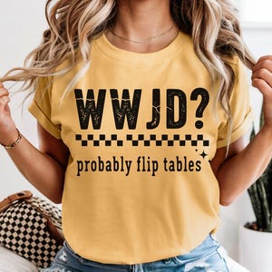 May include: Mustard yellow t-shirt with the text "WWJD? probably flip tables" in black. The shirt has a crew neck and short sleeves. The text is above a black and white checkered line.
