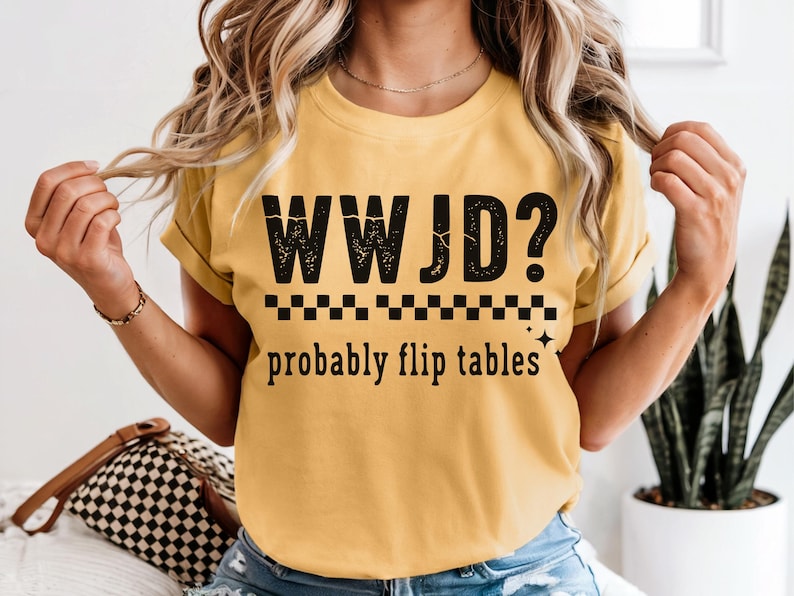 May include: Mustard yellow t-shirt with the text "WWJD? probably flip tables" in black. The shirt has a crew neck and short sleeves. The text is above a black and white checkered line.