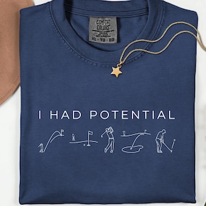 Pode incluir: Camiseta azul marinho com o texto "I HAD POTENTIAL" e ilustrações de golfe em branco. Um colar dourado com um pingente de estrela e um chapéu marrom também são visíveis. A camisa é Comfort Colors XL-TG-EG.