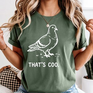 May include: Green t-shirt with a white outline drawing of a pigeon. The text "THAT'S COO" is printed below the bird. The shirt has short sleeves and a crew neck.