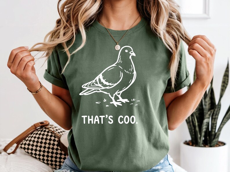 May include: Green t-shirt with a white outline drawing of a pigeon. The text "THAT'S COO" is printed below the bird. The shirt has short sleeves and a crew neck.