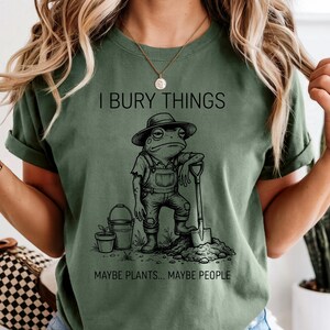 May include: Olive green t-shirt with the text "I BURY THINGS" above a cartoon frog wearing overalls and a hat, leaning on a shovel. Below the frog is the text "MAYBE PLANTS... MAYBE PEOPLE."