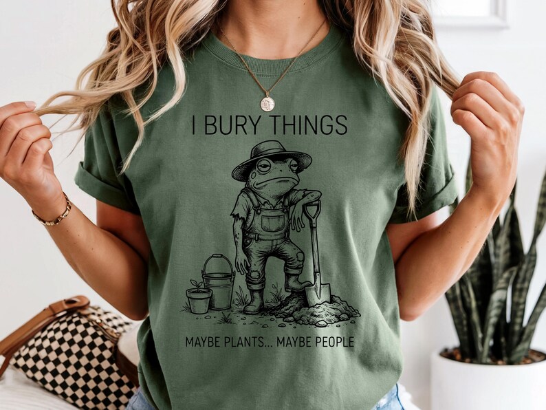May include: Olive green t-shirt with the text "I BURY THINGS" above a cartoon frog wearing overalls and a hat, leaning on a shovel. Below the frog is the text "MAYBE PLANTS... MAYBE PEOPLE."