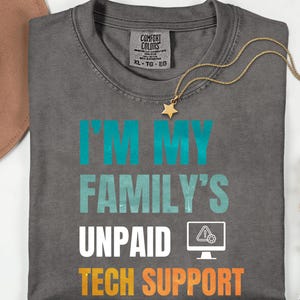 May include: Dark grey t-shirt with the text "I'M MY FAMILY'S UNPAID TECH SUPPORT" in teal, white, and orange. A gold necklace with a star pendant and a brown hat are also visible.
