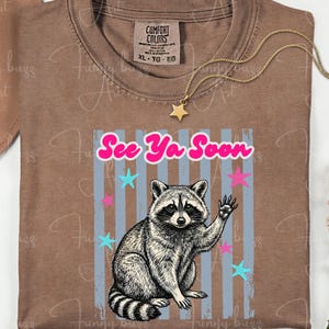Peut inclure: T-shirt marron avec un dessin de raton laveur qui salue, le texte "See Ya Soon" et des &eacute;toiles. Le t-shirt a un col rond et une &eacute;tiquette Comfort Colors.