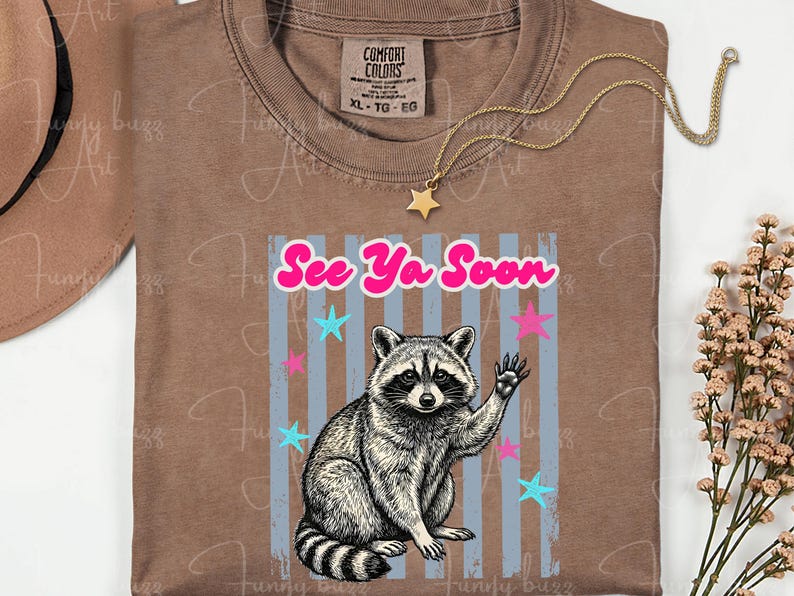 Peut inclure: T-shirt marron avec un dessin de raton laveur qui salue, le texte "See Ya Soon" et des &eacute;toiles. Le t-shirt a un col rond et une &eacute;tiquette Comfort Colors.