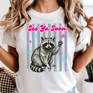 Peut inclure: T-shirt blanc avec une illustration de raton laveur faisant un signe de la main et le texte rose "See Ya Soon". Le motif comprend des &eacute;toiles bleues et roses et des rayures verticales grises, pour un style d&eacute;contract&eacute;.