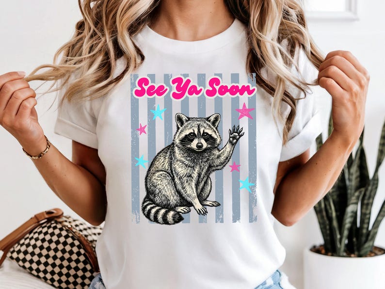 Peut inclure: T-shirt blanc avec une illustration de raton laveur faisant un signe de la main et le texte rose "See Ya Soon". Le motif comprend des &eacute;toiles bleues et roses et des rayures verticales grises, pour un style d&eacute;contract&eacute;.