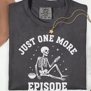 May include: Dark gray t-shirt with the text "JUST ONE MORE EPISODE" and a skeleton graphic holding a remote control. A gold necklace with a star pendant and a bowl of popcorn are also visible. The shirt is folded and has a Comfort Colors tag.