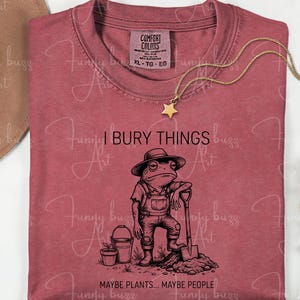 May include: A dusty rose t-shirt with a humorous graphic of a frog in overalls holding a shovel, with the text "I BURY THINGS... MAYBE PLANTS... MAYBE PEOPLE." A gold star pendant necklace and a brown hat are also visible.
