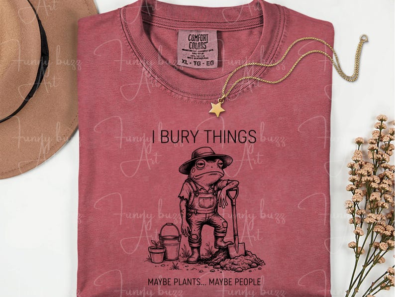 May include: A dusty rose t-shirt with a humorous graphic of a frog in overalls holding a shovel, with the text "I BURY THINGS... MAYBE PLANTS... MAYBE PEOPLE." A gold star pendant necklace and a brown hat are also visible.