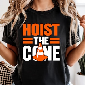 May include: Black t-shirt with the words "HOIST THE CONE" in orange and white. The design includes an orange traffic cone graphic. The shirt is a casual style.