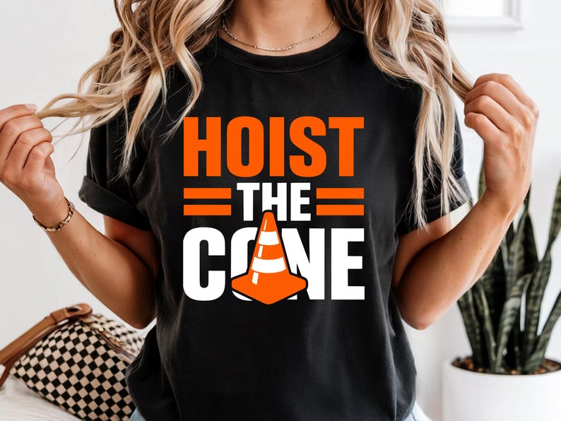 May include: Black t-shirt with the words "HOIST THE CONE" in orange and white. The design includes an orange traffic cone graphic. The shirt is a casual style.