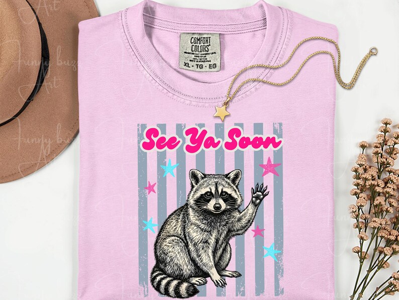 Peut inclure: T-shirt rose clair avec un dessin de raton laveur qui salue. Le texte "See Ya Soon" est en rose au-dessus du raton laveur. Le motif comprend des &eacute;toiles bleues et roses et des rayures verticales grises. Un collier dor&eacute; avec un pendentif &eacute;toile est sur le t-shirt.