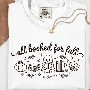 May include: A white t-shirt featuring a black graphic design with the text all booked for fall. The illustration includes a pumpkin, a stack of books, a ghost reading a book, a row of books, a maple leaf, small hearts, and stars.