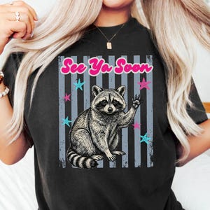 Peut inclure: T-shirt noir avec une illustration de raton laveur qui salue. Le design comprend le texte "See Ya Soon" en rose, avec des &eacute;toiles bleues et roses sur un fond ray&eacute;. Un t-shirt graphique d&eacute;contract&eacute; et amusant.