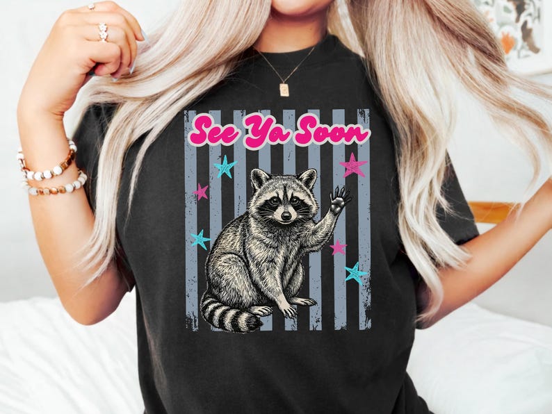 Peut inclure: T-shirt noir avec une illustration de raton laveur qui salue. Le design comprend le texte "See Ya Soon" en rose, avec des &eacute;toiles bleues et roses sur un fond ray&eacute;. Un t-shirt graphique d&eacute;contract&eacute; et amusant.