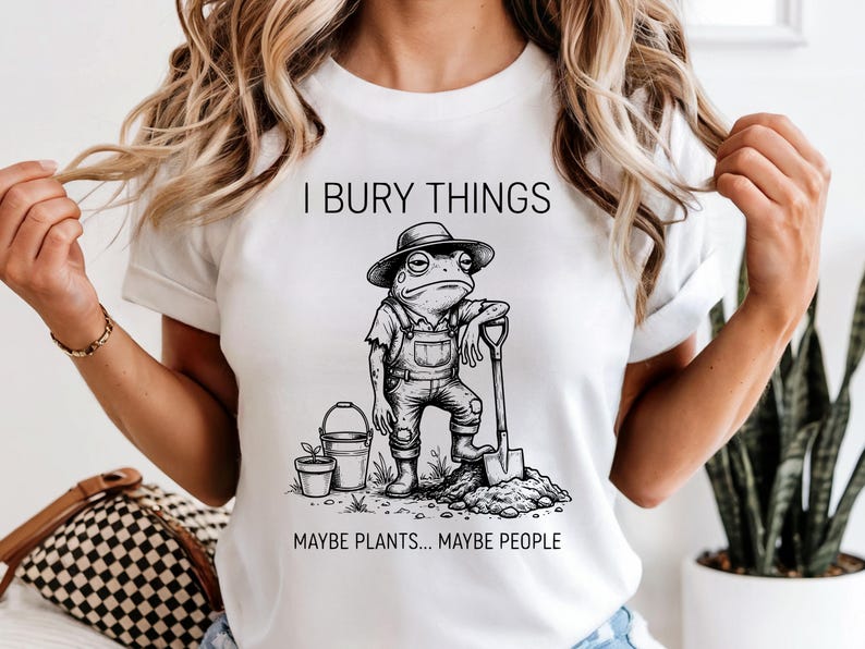 May include: White t-shirt with a humorous graphic of a frog in overalls and a hat, leaning on a shovel. The text reads "I BURY THINGS MAYBE PLANTS... MAYBE PEOPLE."