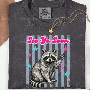 Peut inclure: T-shirt gris fonc&eacute; avec un dessin de raton laveur qui salue, le texte "See Ya Soon" et des &eacute;toiles. Le t-shirt est un Comfort Colors XL-TG-EO. Un collier dor&eacute; avec un pendentif &eacute;toile est pos&eacute; sur le t-shirt.