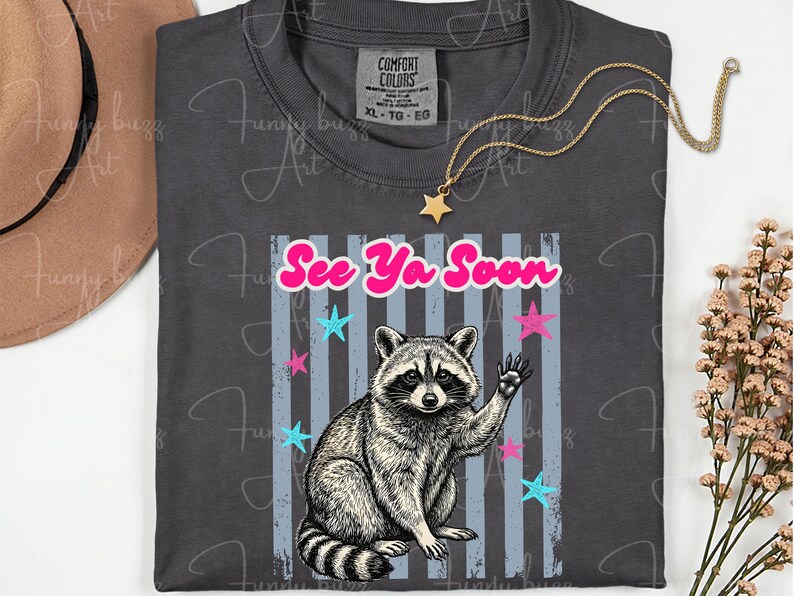 Peut inclure: T-shirt gris fonc&eacute; avec un dessin de raton laveur qui salue, le texte "See Ya Soon" et des &eacute;toiles. Le t-shirt est un Comfort Colors XL-TG-EO. Un collier dor&eacute; avec un pendentif &eacute;toile est pos&eacute; sur le t-shirt.