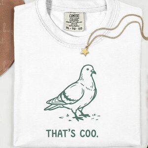 May include: White t-shirt with a green illustration of a pigeon and the text "THAT'S COO." A gold star pendant necklace is draped over the shirt. The shirt has a Comfort Colors tag.