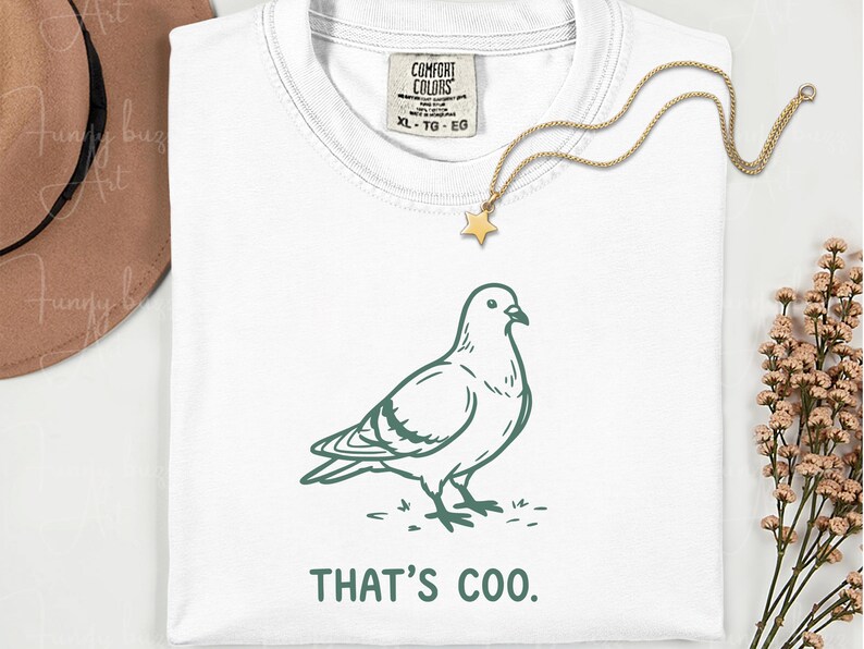 May include: White t-shirt with a green illustration of a pigeon and the text "THAT'S COO." A gold star pendant necklace is draped over the shirt. The shirt has a Comfort Colors tag.