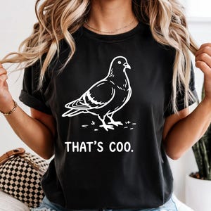 May include: Black t-shirt featuring a white outline of a pigeon with the text "THAT'S COO." The design is a playful take on the bird. The shirt is a casual wear item.