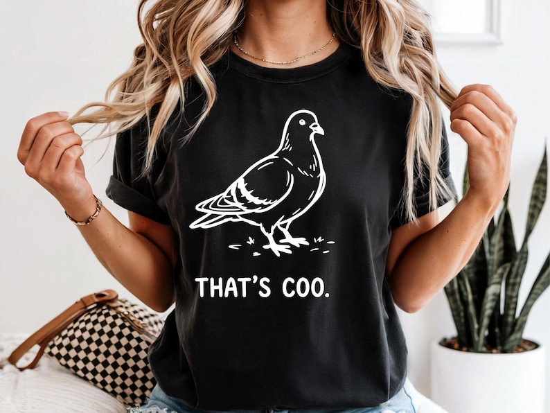 May include: Black t-shirt featuring a white outline of a pigeon with the text "THAT'S COO." The design is a playful take on the bird. The shirt is a casual wear item.