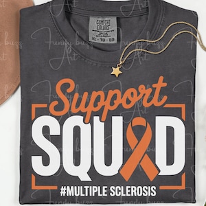 May include: Dark gray t-shirt with the words "Support SQUAD" in white and orange, featuring an orange ribbon. The text "#MULTIPLE SCLEROSIS" is below. A gold star necklace and a hat are also visible.