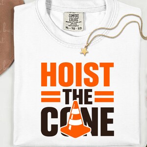 May include: White t-shirt with the words "HOIST THE CONE" in orange and black. The design includes a traffic cone graphic. A gold necklace with a star pendant and a brown hat are also visible.