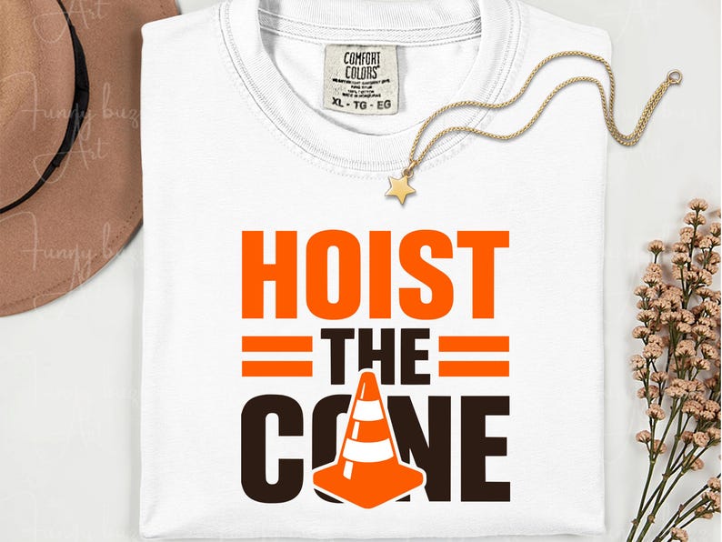 May include: White t-shirt with the words "HOIST THE CONE" in orange and black. The design includes a traffic cone graphic. A gold necklace with a star pendant and a brown hat are also visible.