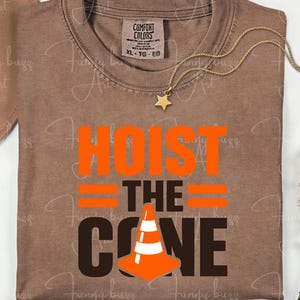 May include: A brown t-shirt with the words "HOIST THE CONE" in orange and brown. The design includes a traffic cone graphic. A gold necklace with a star pendant is draped across the shirt.
