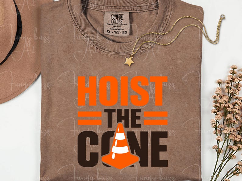 May include: A brown t-shirt with the words "HOIST THE CONE" in orange and brown. The design includes a traffic cone graphic. A gold necklace with a star pendant is draped across the shirt.