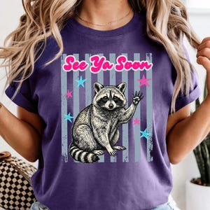 Peut inclure: T-shirt violet avec un dessin de raton laveur qui salue, avec le texte "See Ya Soon" en rose. Le motif comprend des &eacute;toiles bleues et roses et des rayures verticales. Le t-shirt a des manches courtes.