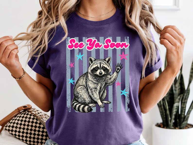Peut inclure: T-shirt violet avec un dessin de raton laveur qui salue, avec le texte "See Ya Soon" en rose. Le motif comprend des &eacute;toiles bleues et roses et des rayures verticales. Le t-shirt a des manches courtes.