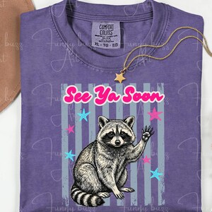 Peut inclure: T-shirt violet avec un dessin de raton laveur qui salue, et l'inscription "See Ya Soon". Le motif comprend des &eacute;toiles roses et bleues et des rayures verticales grises. Un collier dor&eacute; avec un pendentif &eacute;toile est pos&eacute; sur le t-shirt.