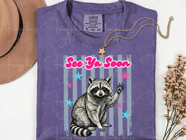 Peut inclure: T-shirt violet avec un dessin de raton laveur qui salue, et l'inscription "See Ya Soon". Le motif comprend des &eacute;toiles roses et bleues et des rayures verticales grises. Un collier dor&eacute; avec un pendentif &eacute;toile est pos&eacute; sur le t-shirt.