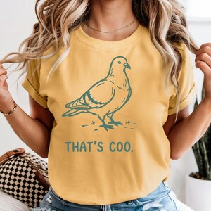 May include: Mustard yellow t-shirt with a teal illustration of a pigeon and the text "THAT'S COO." The shirt has short sleeves and a relaxed fit. The design is simple and graphic, suitable for casual wear.