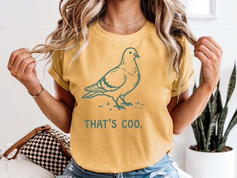 May include: Mustard yellow t-shirt with a teal illustration of a pigeon and the text "THAT'S COO." The shirt has short sleeves and a relaxed fit. The design is simple and graphic, suitable for casual wear.