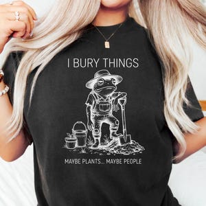 May include: Black t-shirt with a white graphic of a frog in overalls, a hat, and holding a shovel. Text above reads "I BURY THINGS" and below "MAYBE PLANTS... MAYBE PEOPLE". The design is a humorous take on gardening.