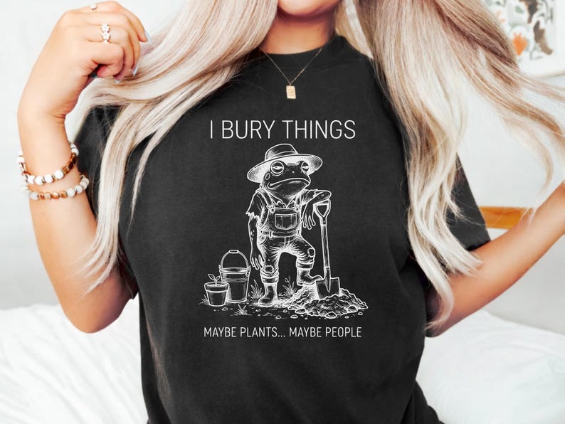 May include: Black t-shirt with a white graphic of a frog in overalls, a hat, and holding a shovel. Text above reads "I BURY THINGS" and below "MAYBE PLANTS... MAYBE PEOPLE". The design is a humorous take on gardening.