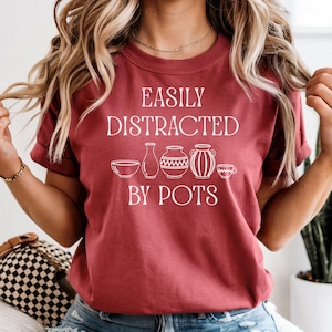 May include: A maroon t-shirt with the text "EASILY DISTRACTED BY POTS" in white. The shirt features illustrations of pottery. The shirt is a short-sleeved crew neck.