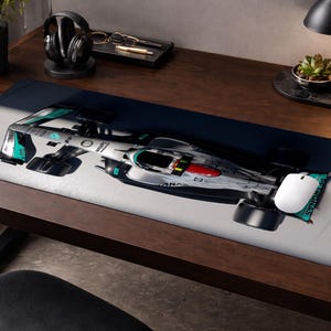 May include: A rectangular mouse pad featuring a Formula 1 race car in silver, black, and teal. The mouse pad has a white mouse on the right side. The desk is made of dark wood.