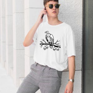 Peut inclure: T-shirt blanc avec un motif noir d'un vautour perché sur une branche avec des crânes. Le design est une illustration monochrome audacieuse. Le t-shirt est de style décontracté à manches courtes.