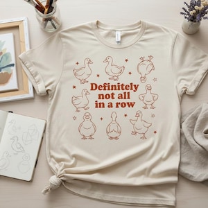 May include: Cream-colored t-shirt with a brown illustration of ducks and the text "Definitely not all in a row." The design features various duck poses and star accents, creating a playful and whimsical aesthetic.