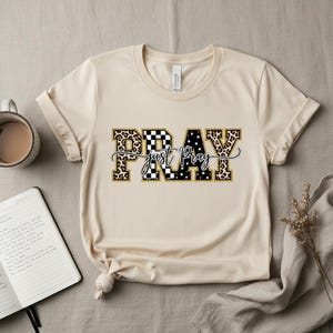 Könnte beinhalten: Ein cremefarbenes T-Shirt mit dem Wort "PRAY" in einer auffälligen, dekorativen Schriftart. Die Buchstaben haben ein Leopardenmuster, ein Schachbrettmuster und ein Sternmuster. Die Worte "just pray" sind in einer kursiven Schrift unter dem Wort "PRAY" geschrieben.