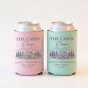 May include: Two can coolers, one pink and one mint green, each with a cabin and mountain graphic. Text on the coolers reads "THE CABIN CREW" and "DANIELLA'S BACHELORETTE - Aspen, Colorado - Est. 2026."